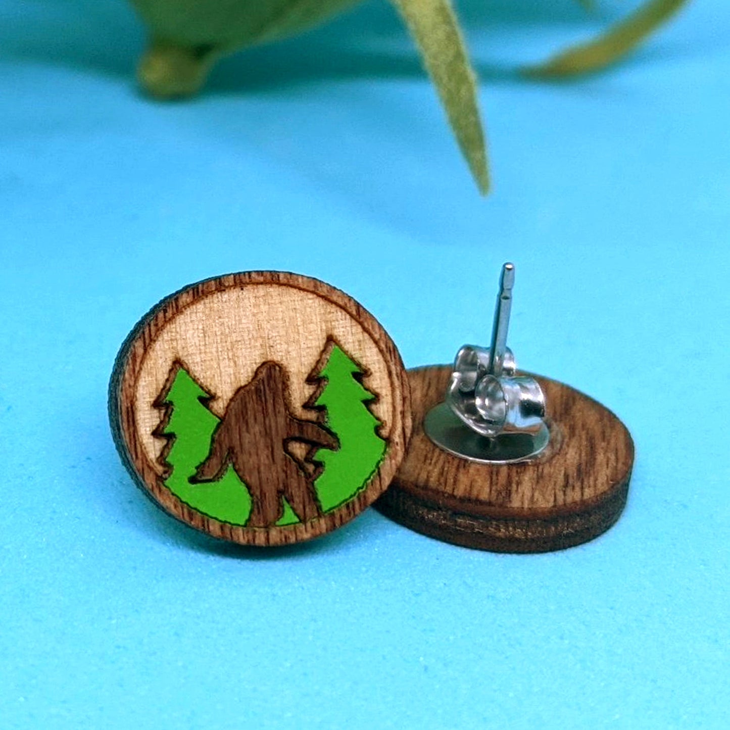 A round wood earring on a blue background. In the center is a carved Bigfoot figure, standing in front of green trees. A second earring is next to it, turned upside down to show the post.