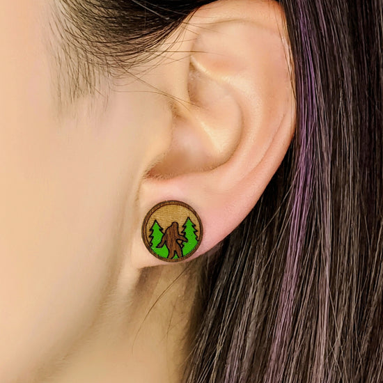 Close up image of a woman’s ear, wearing a round wood earring. In the center is a carved Bigfoot figure, standing in front of green trees.
