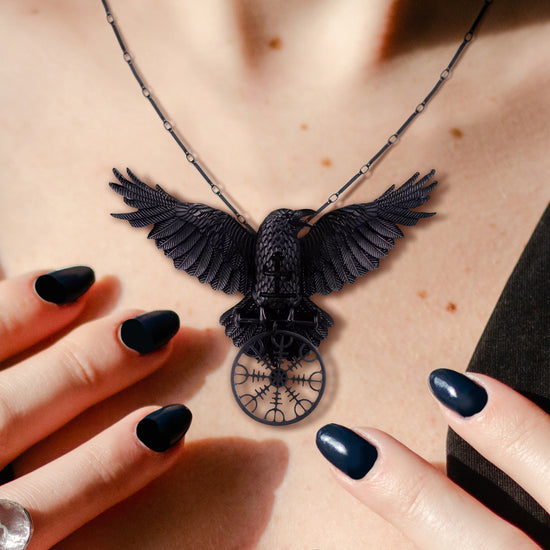 Close up of a black pendant on a black chain, against a model's neck. The pendant is shaped like a black raven with its wings outstretched. The raven is perched on a round Norse-style compass.