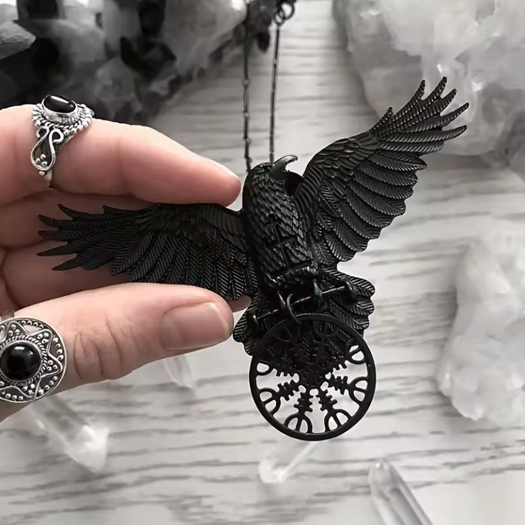 Close up of a black pendant on a black chain, held between a model's fingers. The pendant is shaped like a black raven with its wings outstretched. The raven is perched on a round Norse-style compass. In the background is a light grey wooden table with crystals strewn about the surface.