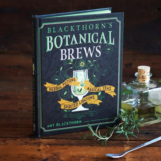 Load image into Gallery viewer, A black book on a wood table. A green and white border runs along the edges. At the top in green and white text is &amp;quot;blackthorn&amp;#39;s botanical brews.&amp;quot; In the center is a drawing of a white goblet with green liquid in in, with green vines surrounding it. At the sides of the goblet are yellow banners, with black text saying &amp;quot;herbal potions, magical teas, spirited libations.&amp;quot; Next to the book is a glass vial, a sprig of herbs, and a metal cocktail stirrer.
