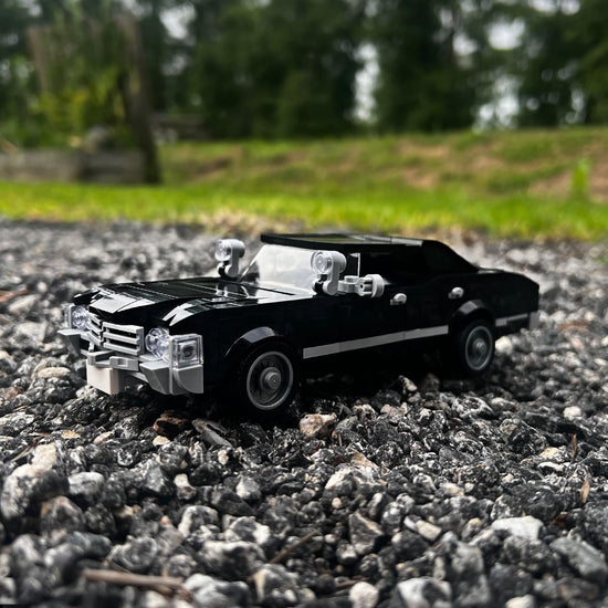 A model of a black 1967 Chevy Impala, built with Lego bricks, parked on small rocks in a field. 