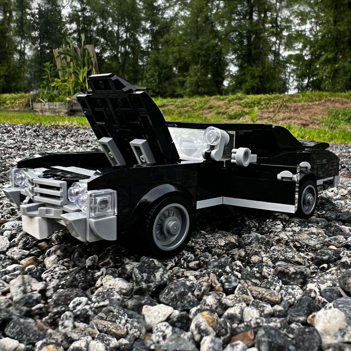 A model of a black 1967 Chevy Impala, built with Lego bricks, parked on small rocks in a field.  The hood and doors are opened on the model.