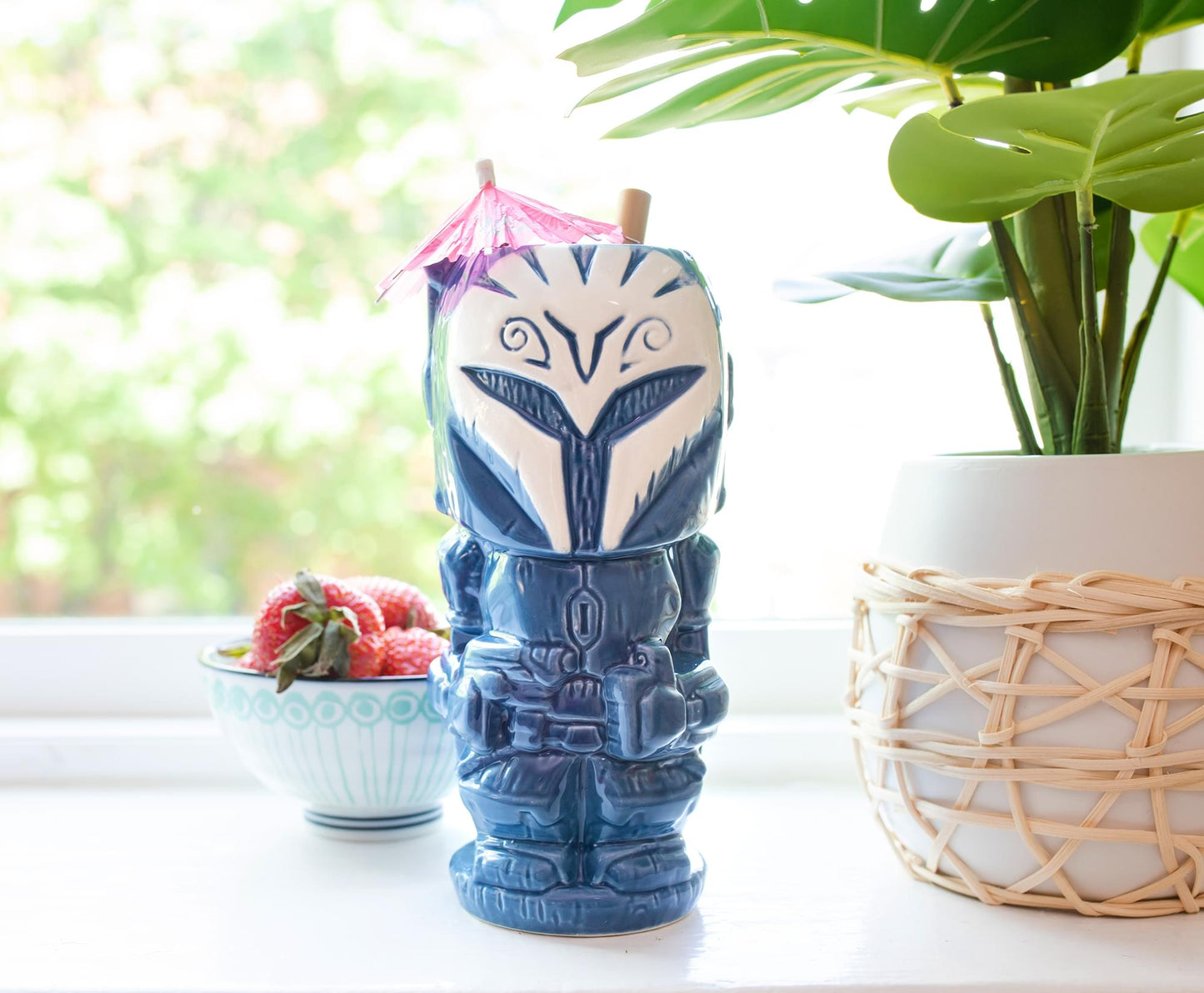 A blue tiki mug depicting the Mandalorian character Bo Katan. The mug is on a table next to a bowl of strawberries and a potted plant.