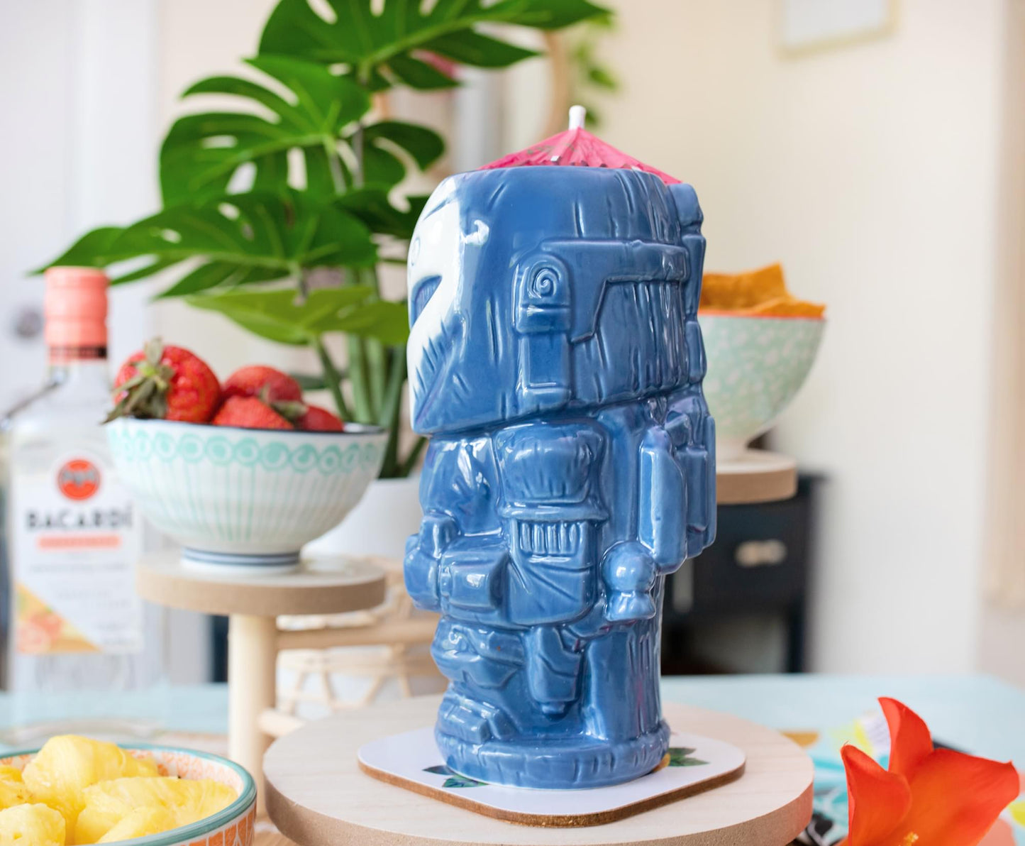 A blue tiki mug depicting the Mandalorian character Bo Katan. The mug is on a table next to bowls of fruit and a potted plant.
