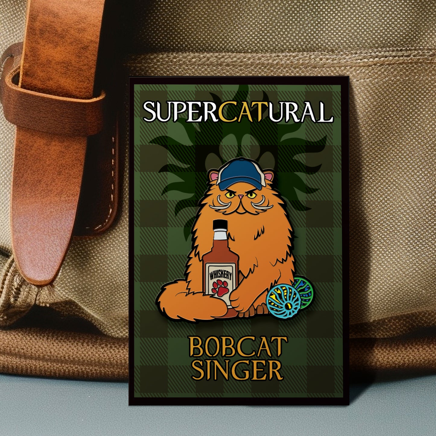 An enamel pin with a green plaid background, against a tan canvas messenger bag. In the center of the pin is a cartoon drawing of an orange bobcat wearing a blue trucker hat, with a whiskey bottle and two cat toys in front of him.  At the top and bottom is white and yellow text saying SuperCatural: Bobcat Singer." In the pin's background is a black paw print, reminiscent of the anti-possession symbol