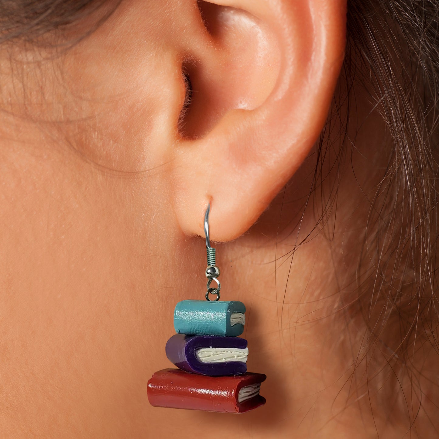 A close-up of a woman's ear. She is wearing a pair of small, clay earrings in the shape a stack of books. The earrings are on a set of silver earring hooks