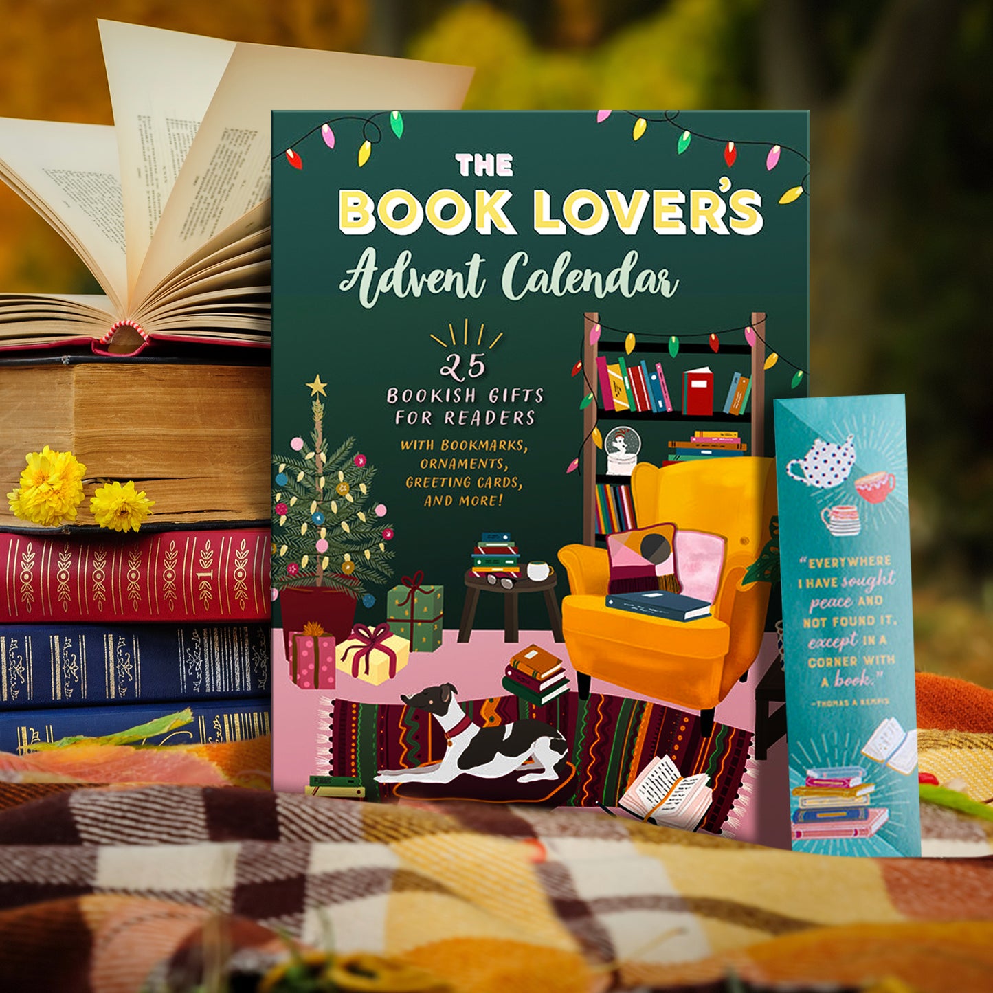 A green book on a plaid blanket, next to a stack of books.. At the top of the cover is pink, green, and yellow text saying "The Book Lover's Advent Calendar," against a green background. At the middle and bottom of the cover is a drawing of a living room decorated for Christmas. Books are piled on a yellow chair, the floor, on a brown bookcase, and on a small table. A dog lays on a pillow on the floor.