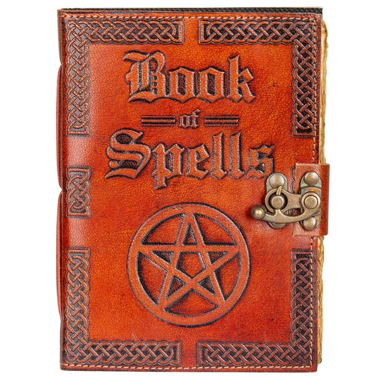 A brown leather book on a wooden table. Embossed text on the front cover says "book of spells," with an embossed pentagram symbol beneath it. On the right edge is a metal clasp to keep the book closed.