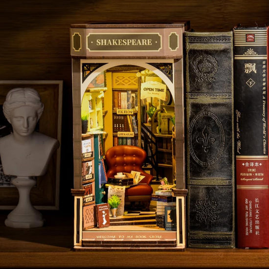 Close up of a wooden book nook light, depicting Shakespeare's Bookstore in France, on a shelf. The interior is filled with tiny books on shelves, surrounding a red leather armchair and a table with a miniature teacup and saucer on it. On the front of the model is a black sign with "Shakespeare" in gold text. 