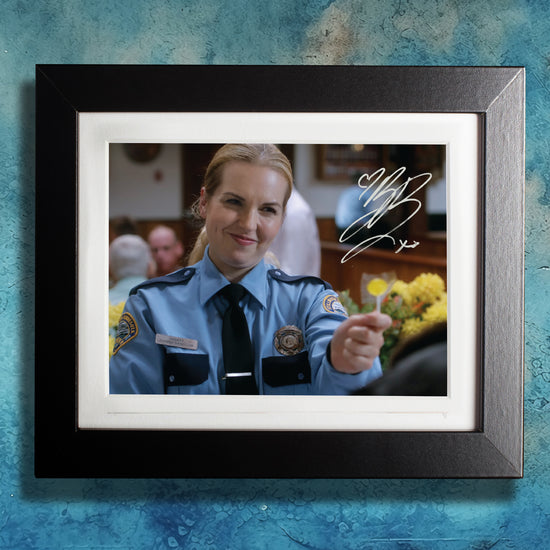 A black framed photograph against a blue wall. The photograph depicts actor Briana Buckmaster, dressed as Deputy Donna Hanscum from the series "Supernatural." Briana is in a blue police uniform, holding a yellow lollipop. In the background is a diner. On the top right side is Briana's autograph in white ink.