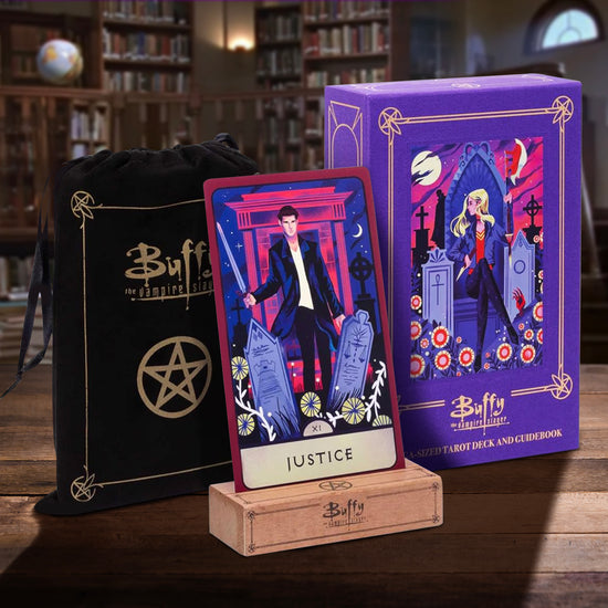 Close up of a purple box, a giant tarot card, and a black felt bag, on a wooden table, set inside the library from the TV series "Buffy The Vampire Slayer." The box top depicts a cartoon drawing of Buffy sitting on a stone throne in a graveyard. The tarot card depicts a drawing of Angel wielding a sword. The black bag has the Buffy logo on the front in gold ink.