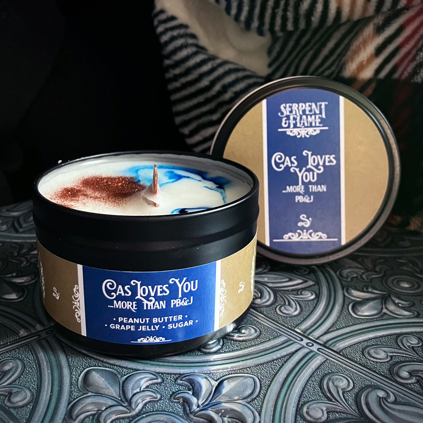 A black candle tin containing a white candle with a blue swirl and copper candle-safe glitter. The blue-and-tan label reads "Cas Loves You...More Than PB&J. Peanut butter / grape jelly / sugar"