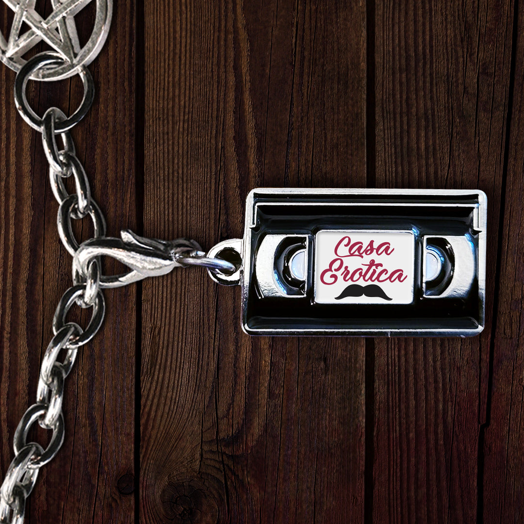 Close up of an enamel charm on a silver chain, against a dark wood table. The charm is shaped like a VHS tape, with a white label on the front. Red text says "cana erotica." A black mustache is drawn under the text.