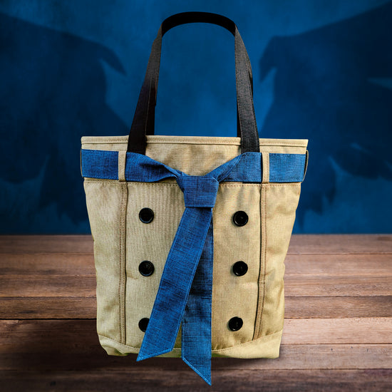 Load image into Gallery viewer, A khaki-colored tote bag with a blue necktie around the top, sitting on a wooden table. In the background is a blue wall with a shadow of angel wings behind the bag.
