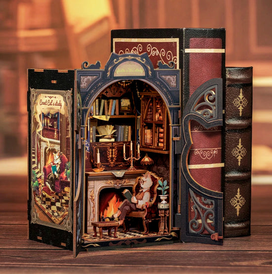 A paper book nook shelf decoration on a wooden table, next to a pair of hardcover books. The front of the book nook has ornate green doors, with a cat's head shaped cut out in the center. Inside is a miniature library, with a cat sitting on a plush chair, reading a book.