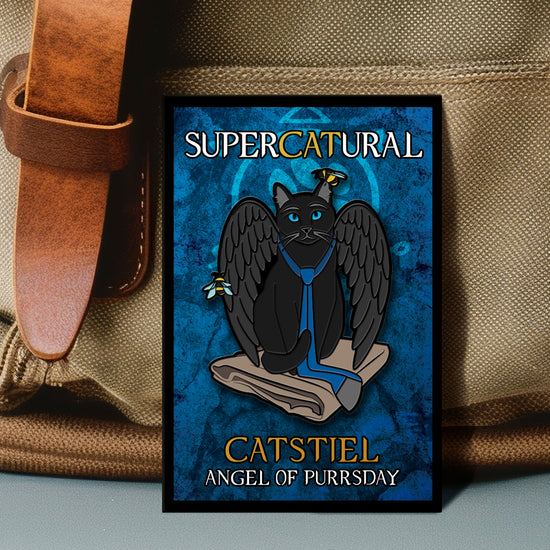 A rectangular enamel pin against a tan canvas bag. The pin depicts a black cat with black wings, sitting on a folded tan raincoat. A blue necktie is around the cat's neck, facing backward. Behind the cat is a blue background with a light blue paw print. White and yellow text says "Supercatural. Catstiel, angel of purrsday."