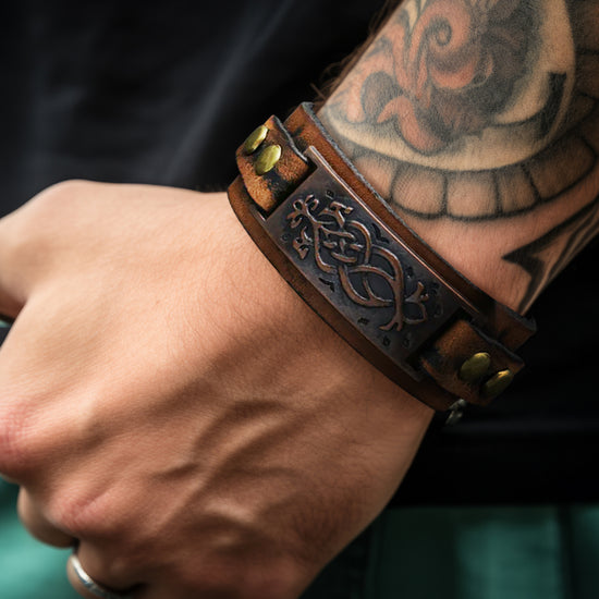 Close up of a model's wrist, wearing a dark brown leather cuff bracelet inspired by Viking imagery/style. It has brassy metal details, an adjustable button closure, and a brown metal plate with visual detailing. A large tattoo adorns the model's wrist.