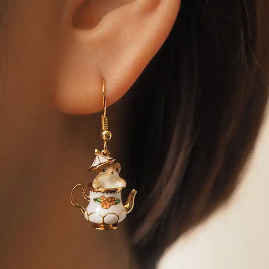 Close up view of an earring hanging from a model's ear. The earring is shaped like an old-fashioned teapot, with a red flower in the center and gold accenting. The teapot's lid is ajar, with a tiny chipmunk peeking out.