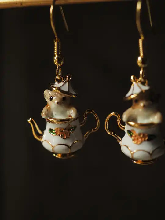 Close up view of two earrings against a black background. The earrings are shaped like an old-fashioned teapot, with a red flower in the center and gold accenting. The teapot's lid is ajar, with a tiny chipmunk peeking out.