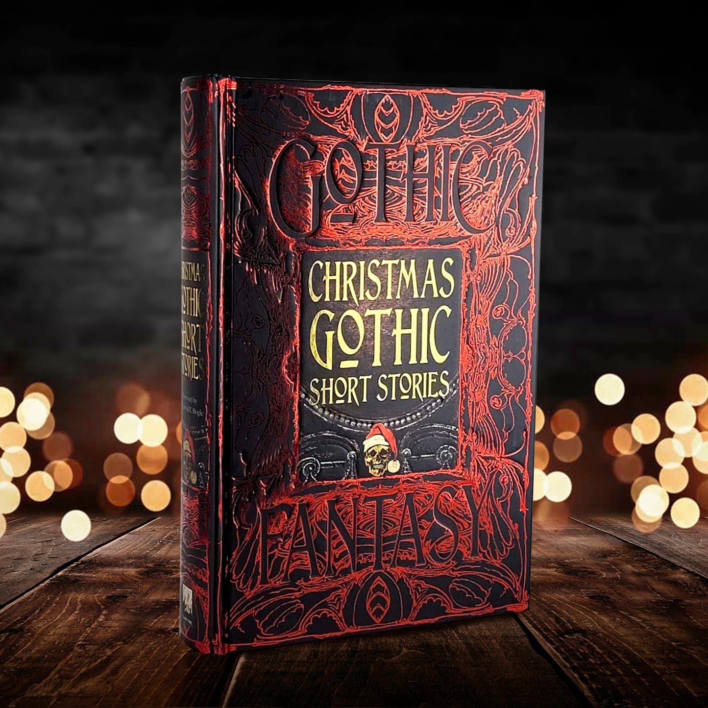 A red and black book on a wooden floor. The background of the cover depicts a gothic floral pattern in red against black, with the words gothic fantasy spelled out in the pattern. In the center is a gray stone tablet, with a skull at the bottom wearing a Santa Claus hat. Yellow text above the skull says Christmas Gothic Short Stories. Behind the book are tiny white lights, against a smoky gray background.