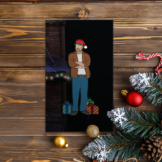 An enamel pin depicting Bobby Singers  in a  Santa hat, arms  crossed, standing next to  Christmas gifts