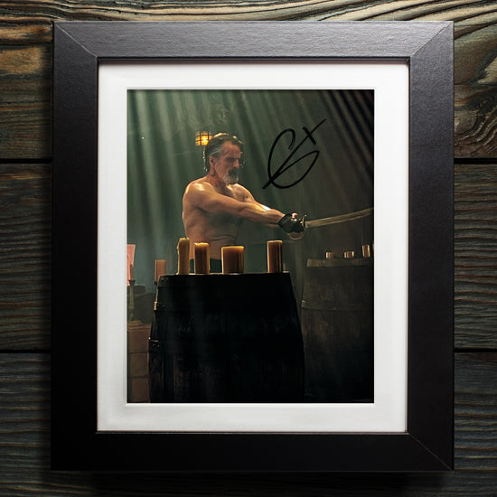 A black framed photograph against a blue wall. The photograph depicts actor Con O'Neill, portraying the character Izzy Hands in "Our Flag Means Death." He is holding a cutlass in his hands, and is standing shirtless in front of a barrel with light candles on top of it. In the top right corner is Con's autograph in black ink.