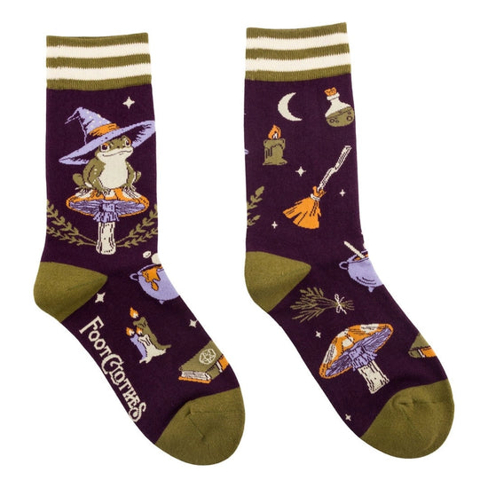 A pair of crew length purple socks. The socks feature a pattern of mushrooms, candles, spell books, and other witch-themed items. On the side is a green toad wearing a purple witch hat, sittong on an orange mushroom.