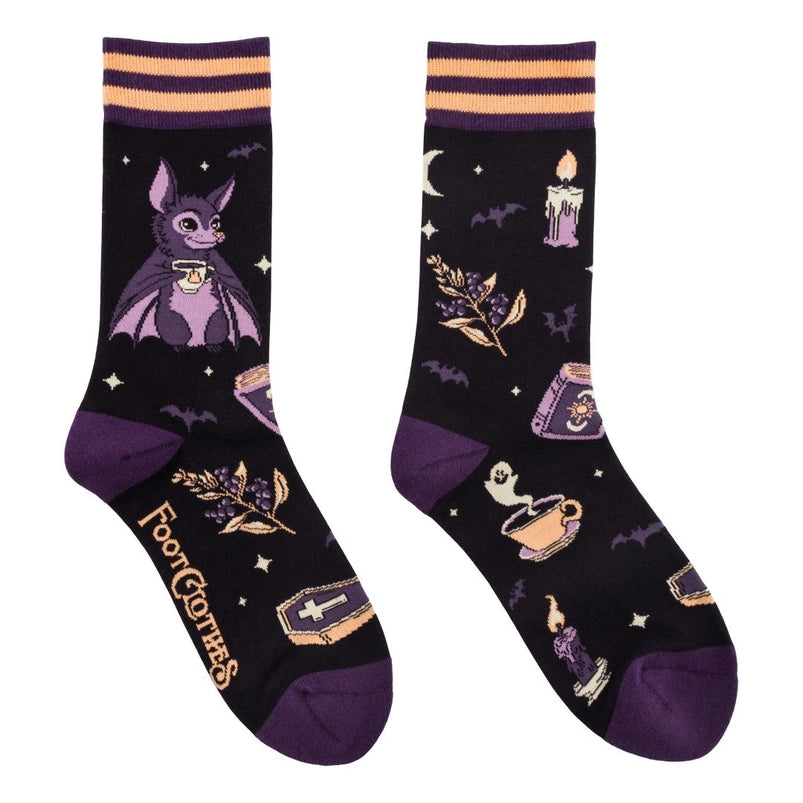 A pair of crew length black socks. The socks feature a pattern of candles, spell books, coffins, and other spooky items. On the side is a purple bat holding a white tea cup.