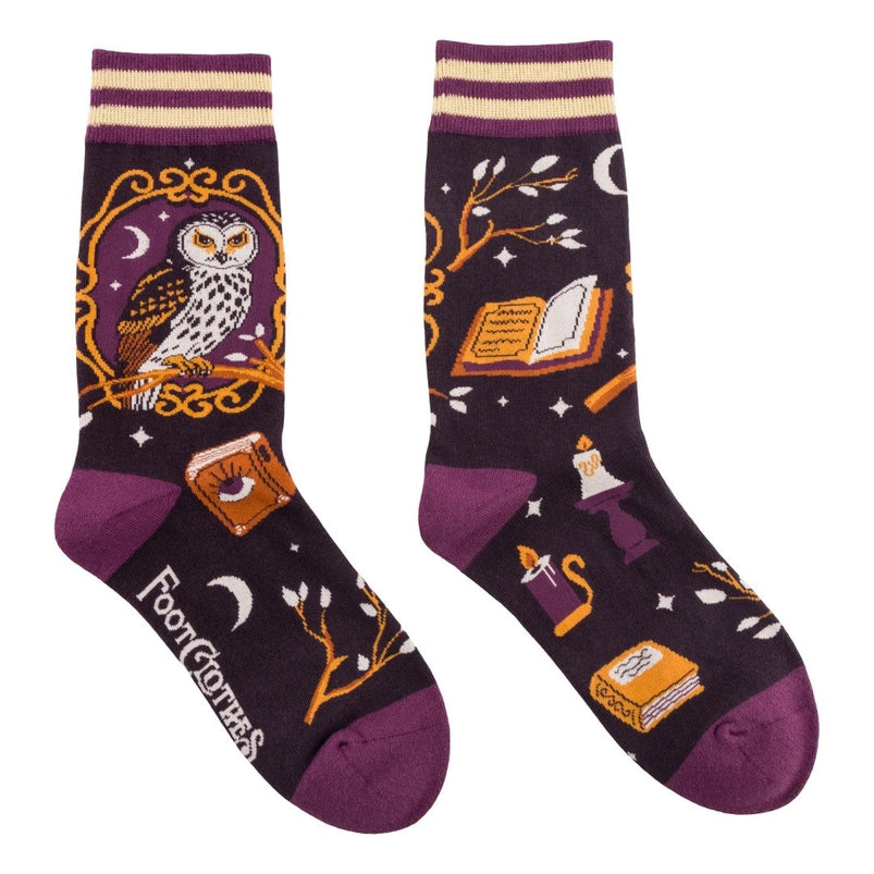 A pair of crew length black socks. The socks feature a pattern of branches, spell books, moons, and other spooky items. On the side is a white and black owl on a yellow perch.