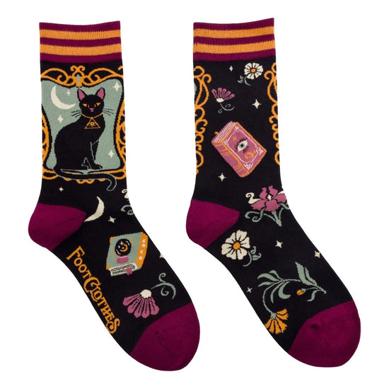 A pair of crew length black socks. The socks feature a pattern of flowers, spell books, and other spooky items. On the side is a black cat against a green square.