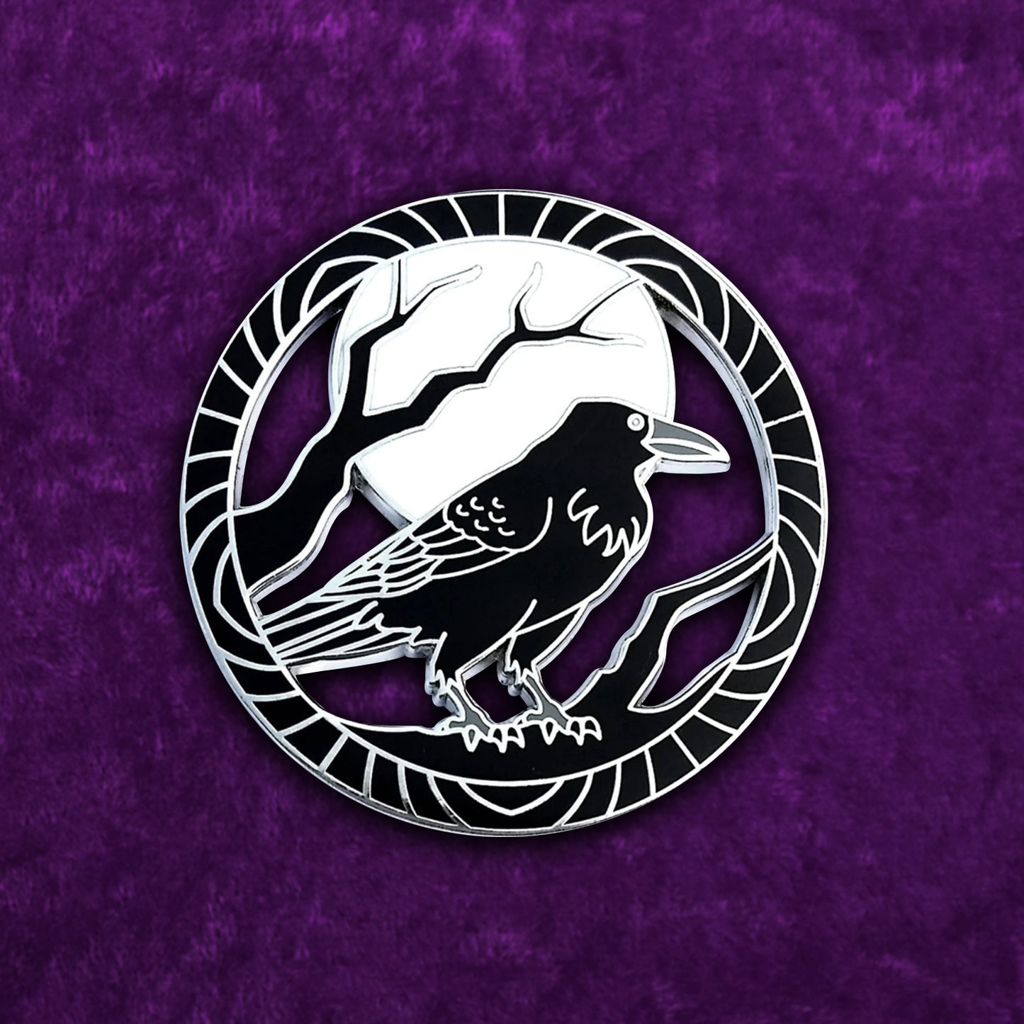 Close up view of a black and silver pin on a white background. The pin depicts a black raven perched on tree branches under a full moon, inside a circle. The pin is. set against a velvety, dark purple background.