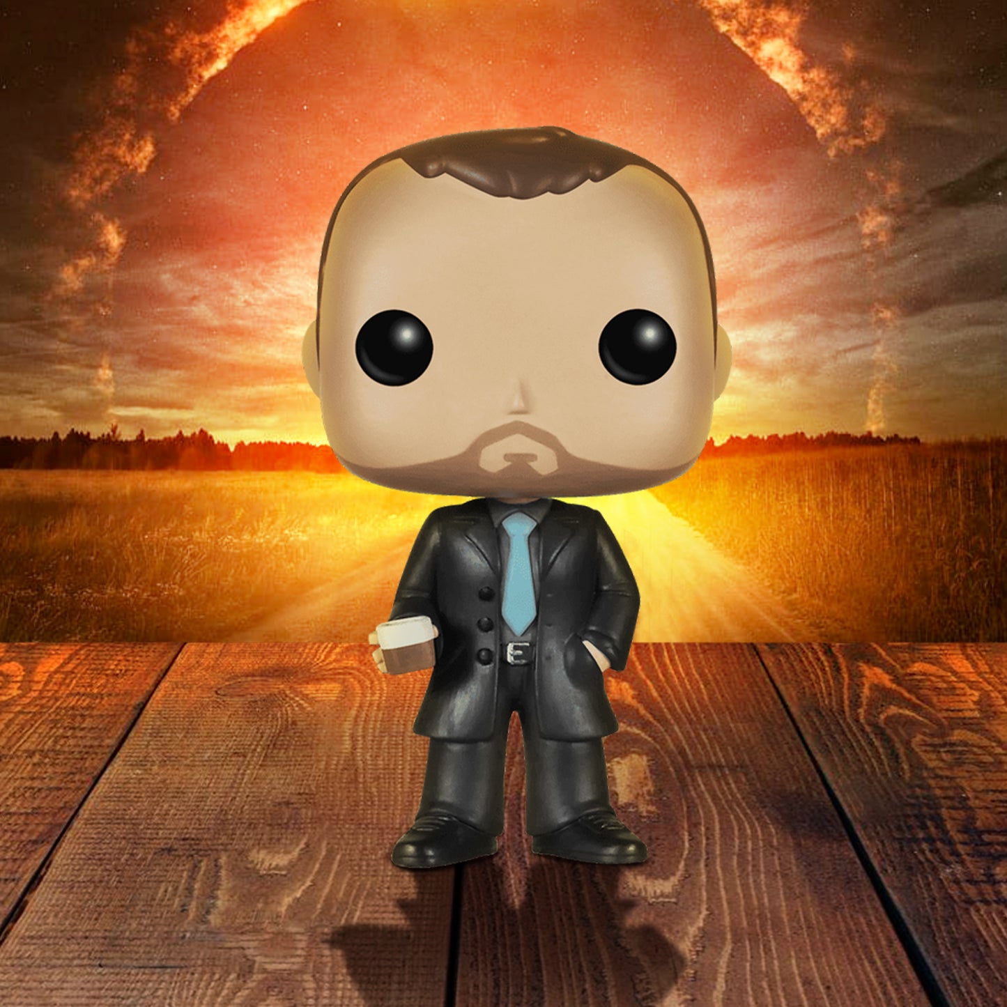 A vinyl Funko Pop figure of the character Crowley from the TV series "Supernatural," standing on a wooden deck. The figure is in a black suit with a light blue tie. In his right hand is a glass depicting single malt whiskey. In the background is a circle of fire in the sky, above a field.