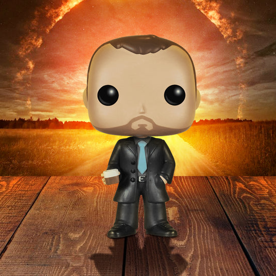A vinyl Funko Pop figure of the character Crowley from the TV series "Supernatural," standing on a wooden deck. The figure is in a black suit with a light blue tie. In his right hand is a glass depicting single malt whiskey. In the background is a circle of fire in the sky, above a field.