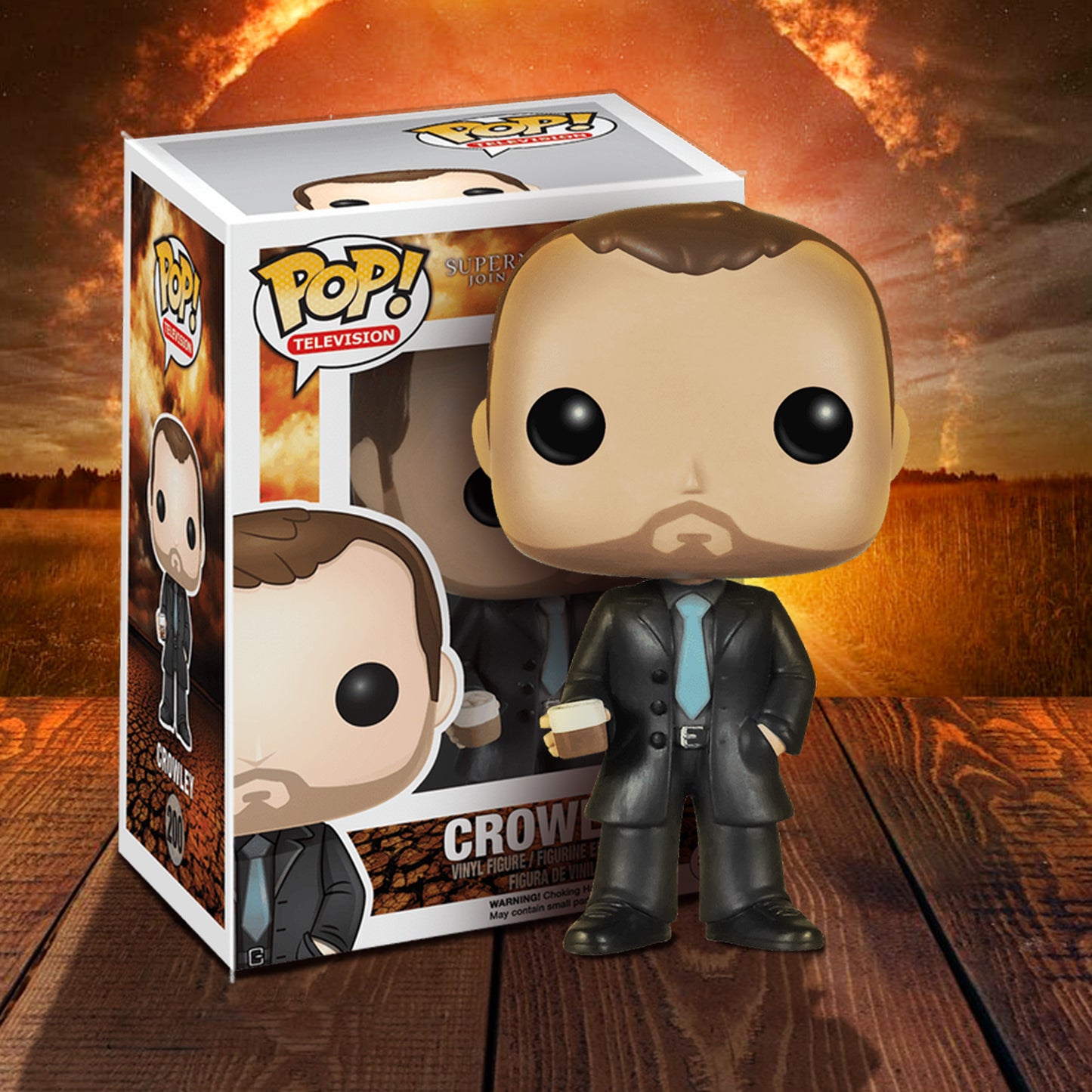 A vinyl Funko Pop figure of the character Crowley from the TV series "Supernatural," on a wooden deck. The figure is in a black suit with a light blue tie. In his right hand is a glass depicting single malt whiskey. Next to the figure is its box. In the background is a circle of fire in the sky, above a field.