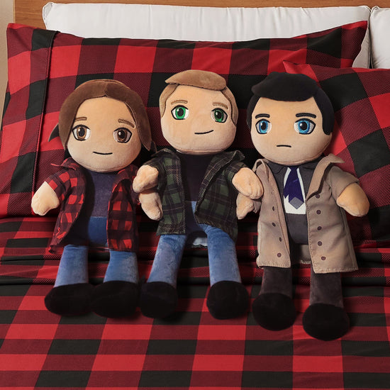 Three plush toys depicting SPN characters sitting on a red and black flannel bedspread against flannel pillows.