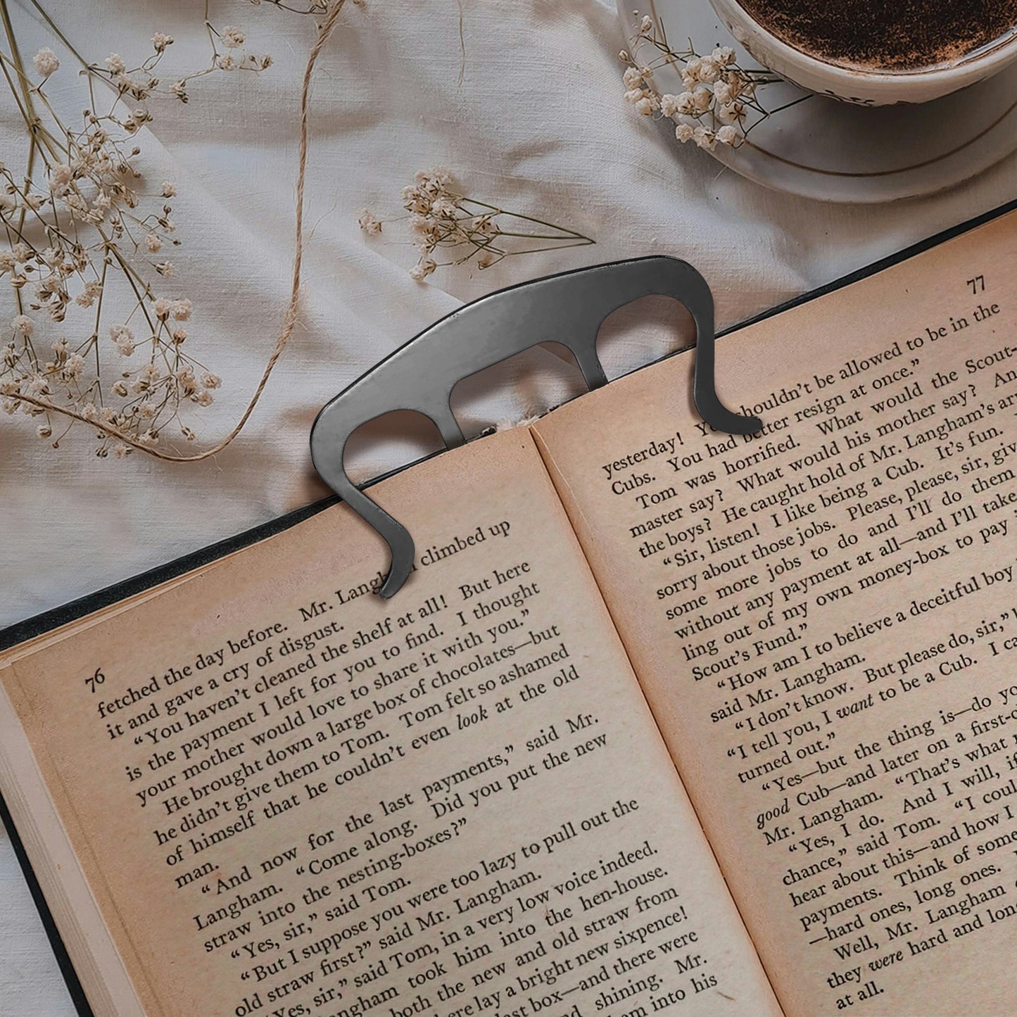 An open hardcover book on a white tablecloth. Inserted into the top is a four-pronged silver metal clip, holding the book open at its current page. Next to the book is a white coffee cup on a saucer, with baby's breath flowers strewn about.