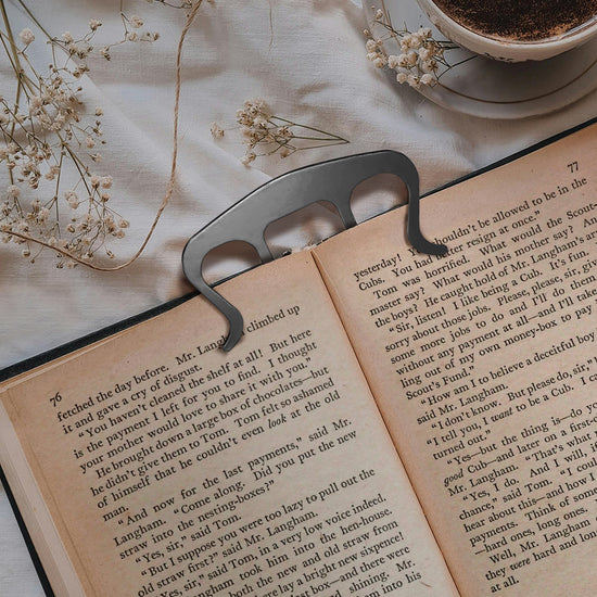 An open hardcover book on a white tablecloth. Inserted into the top is a four-pronged silver metal clip, holding the book open at its current page. Next to the book is a white coffee cup on a saucer, with baby's breath flowers strewn about.