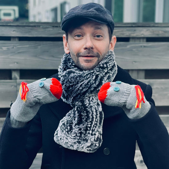 An image of actor DJ Qualls in a winter coat and scarf. On his hands are a set of knit grey flip-top mittens, each with two blue button 'eyes' sewn onto the flip top. Red yarn forms a sock puppet 'mouth' at the top and there is red-and-yellow yarn 'hair' sewn below the knuckles. 