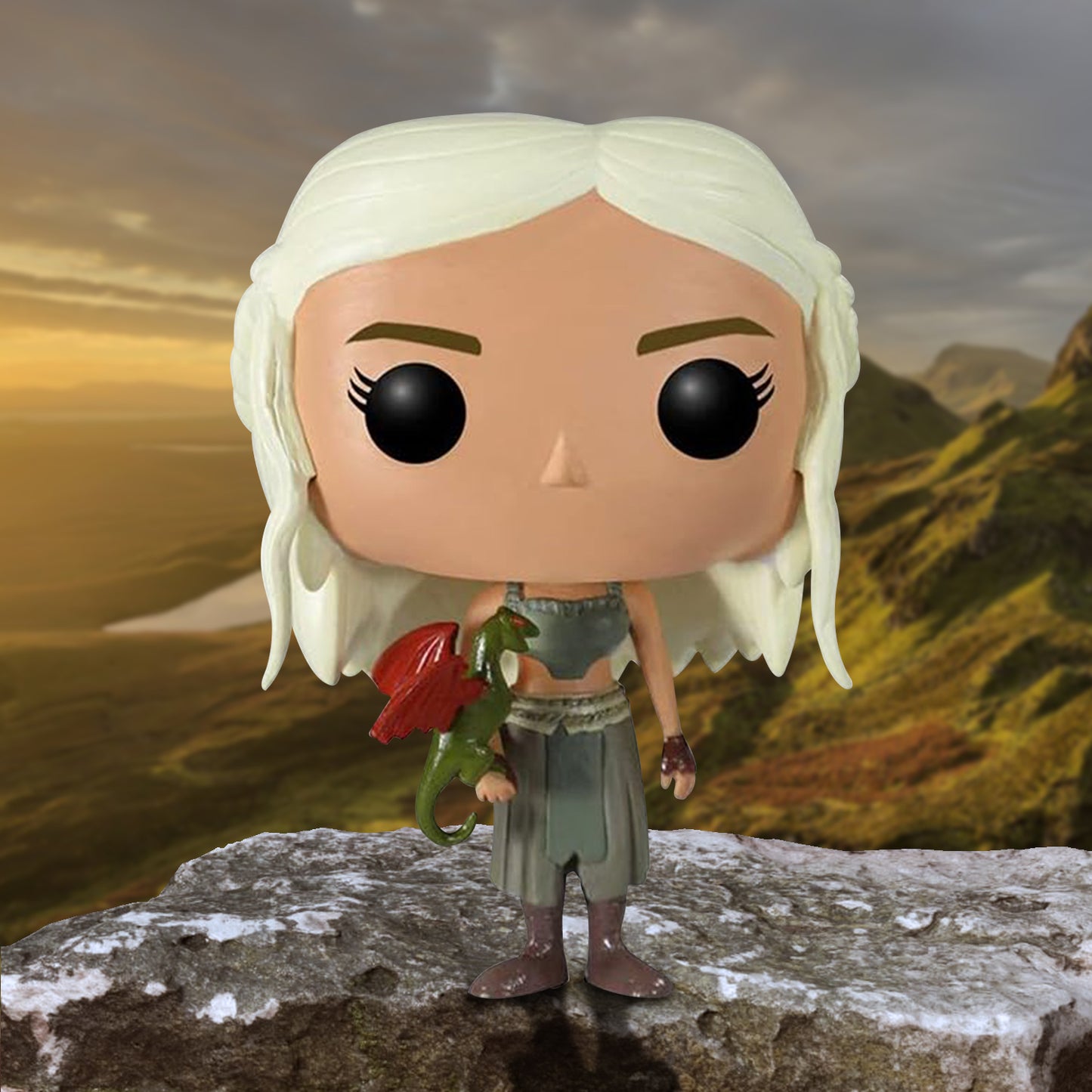 A bobblehead figure depicting Daenerys Targaryen in a green outfit, with a red dragon curled on her hand. She is standing on a rock ledge, with mountains in the background.