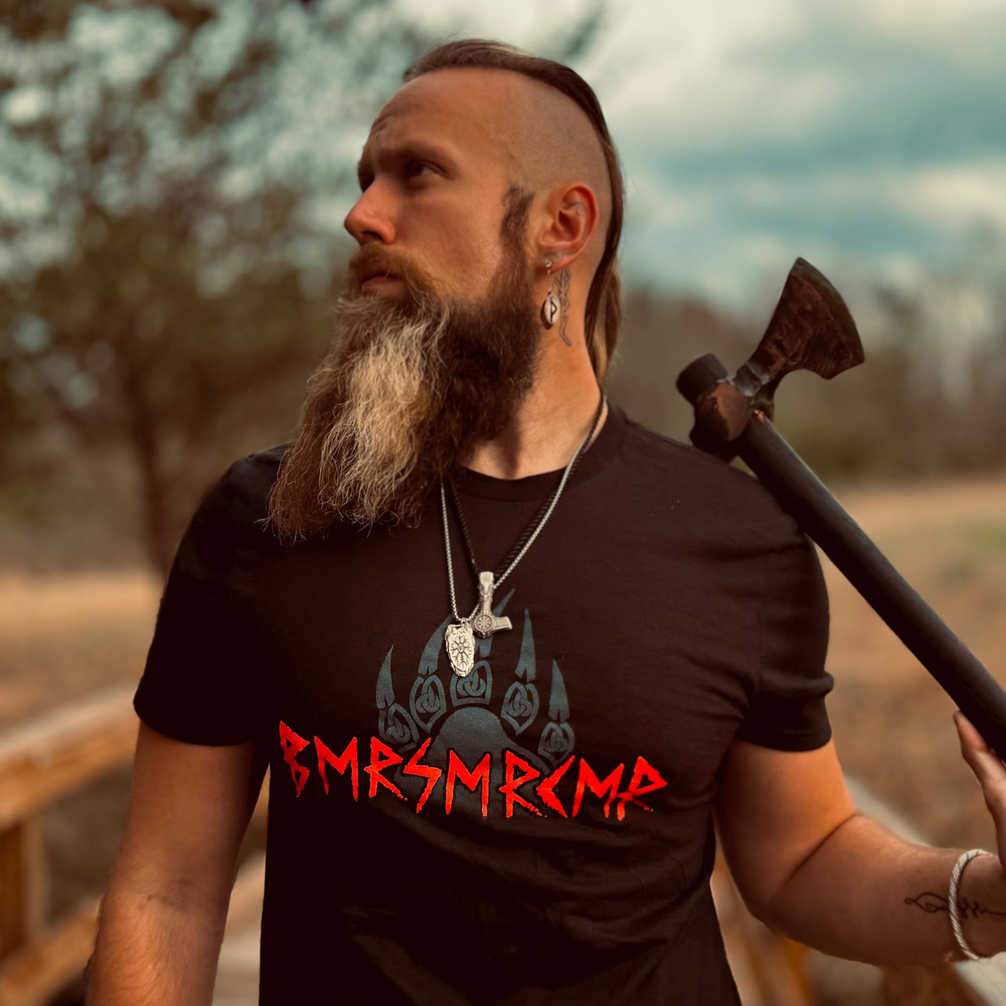 A photo of David Christiansen in a black crew neck tee, holding an axe over one shoulder. The shirt has a blue bear claw behind bold red, carving-style letters that visually resemble "BMRSMRCMR", but translate to "BERSERKER".