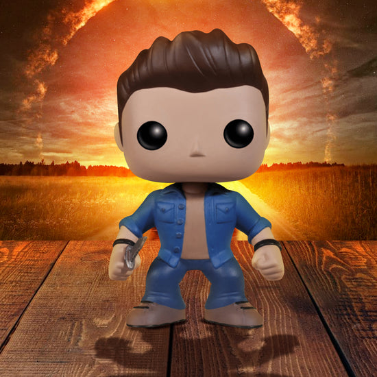 A vinyl Funko Pop figure of the character Dean Winchester from the TV series "Supernatural." The figure is in blue jeans and a blue denim shirt, and is carrying a knife in one hand. The figure is standing on a wooden deck, with a fiery sky in the background