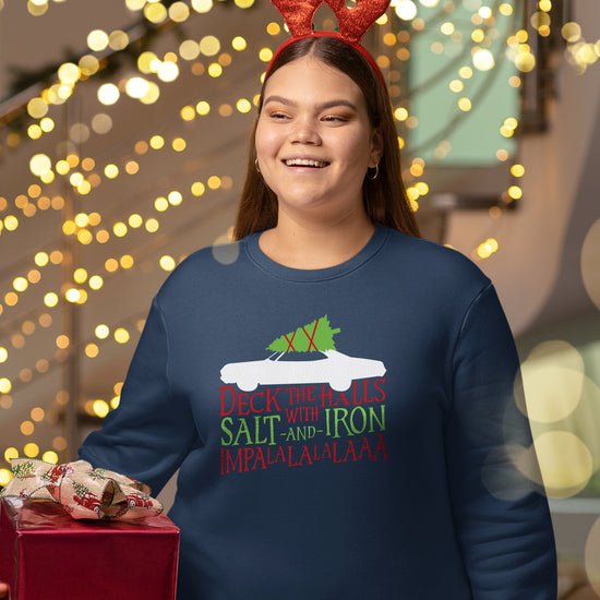 A woman wearing a navy blue crewneck. The crewneck features a while silhouette of a vintage Impala with a green Christmas tree tied to the roof. The crewneck reads "Deck the halls with salt and iron - Impala-la-la-la-laaa" in red and green font below the car.