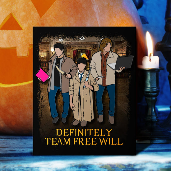 A pin depicting the three main characters from the TV series "Supernatural," but with the actors dressed in each others' costumes. In the center is Jared as Castiel, on his knees to appear shorter. Yellow text at the bottom says "Definitely team free will." Behind them is a jack-o-lantern next to a burning candle.