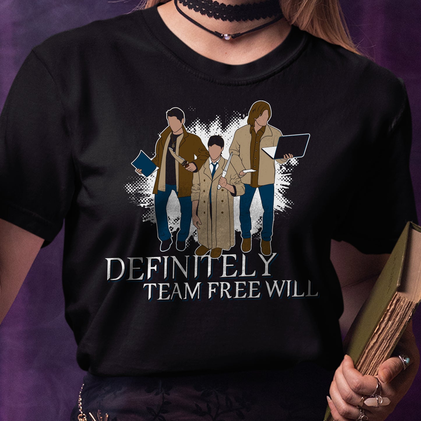 A female model in a black tee shirt against a purple background. On the shirt is the famous behind the scenes picture of Misha, Jared, and Jensen trading their characters's outfits. White text underneath says "Definitely Team Free Will."