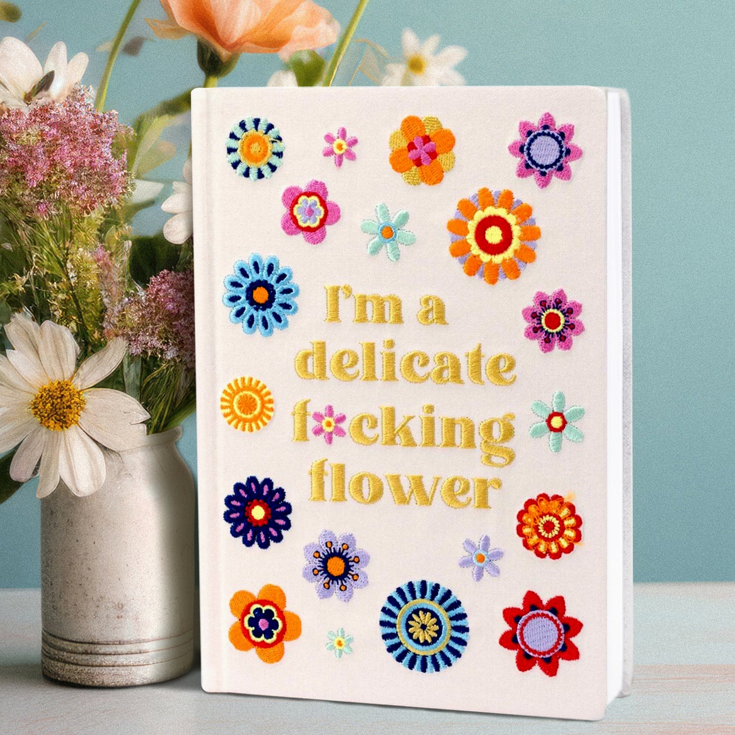 A white hardcover book on a white table, next to a ceramic vase filled with flowers. On the cover of the book is yellow embroidered text saying "I'm a delicate f*cking flower." Around the text are multi-colored embroidered flowers. A light blue background is behind the book.