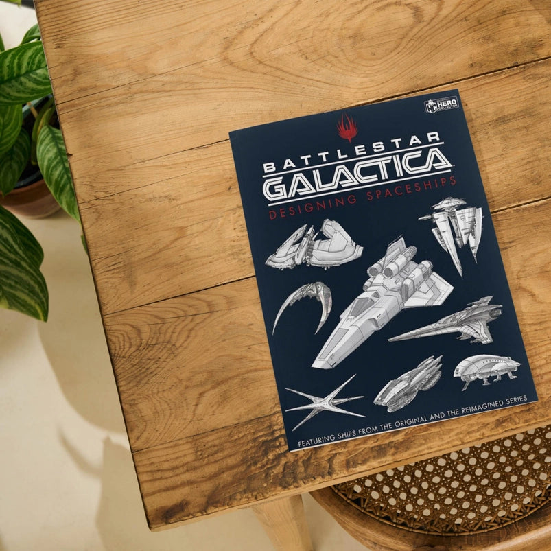 A black book on a wooden table depicting pencil drawings of various spaceships from the Battlestar Galactic series. Red and white text says Battlestar Galactica, Designing Spaceships.