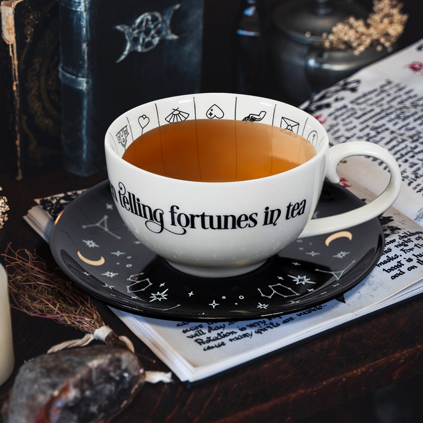 A white teacup with a black saucer, sitting on a book page. around the cup are books, herbs, and other items used in spellcasting and fortune telling.