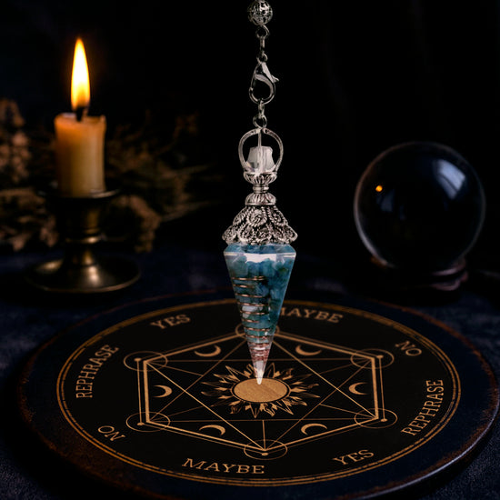 A cone-shaped blue crystal pendulum on a silver chain, hanging over an oracle plate on a table. In the background are a burning candle and a crystal ball.