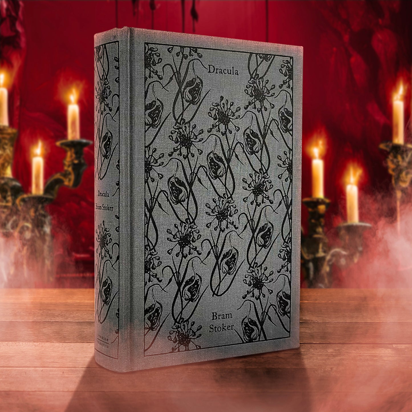 A copy of the Penguin Clothbound edition of DRACULA by Bram Stoker, standing on a dark wood table in front of a dark red wall and vintage-style candles.. The cloth covering is a stone-grey and there's an all-over pattern on the front.